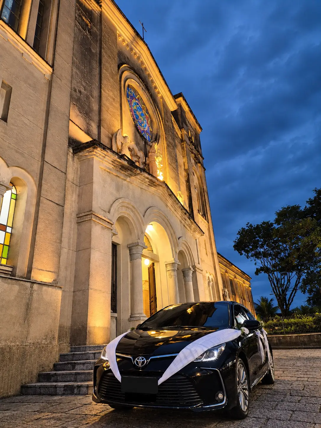 Black sedan decorated with a white wedding ribbon in front of a church lit up at nightfall