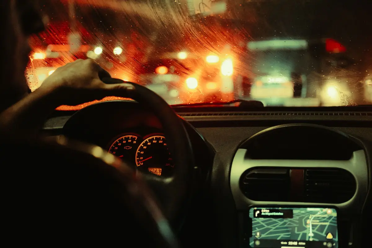 Hand on the steering wheel of a car at night in São Paulo, with the dashboard and GPS lit and blurred traffic through the windshield
