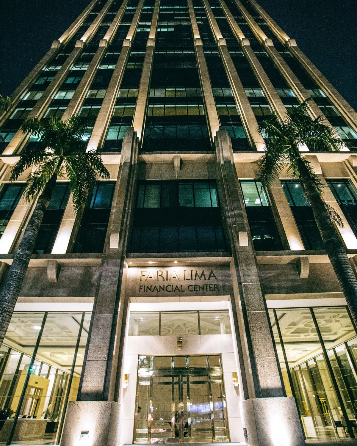 Façade of the Faria Lima Financial Center lit up at night in São Paulo, with palm trees at the entrance
